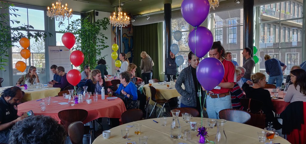 Bezoekers lancering Rotterdammer Centraal, meerdere mensen zijn staand aan of staand tussen grote ronde tafels met elkaar in gesprek over de onderwerpen die op kaartjes zijn geschreven. Op de tafels staan gekleurde balonnen gevuld met helium aan een gewichtje. Dit alles in het historisch gebouw van de Machinist, een voormalige school voor technische scholieren opgeleverd in 1952 met een industrieel design.