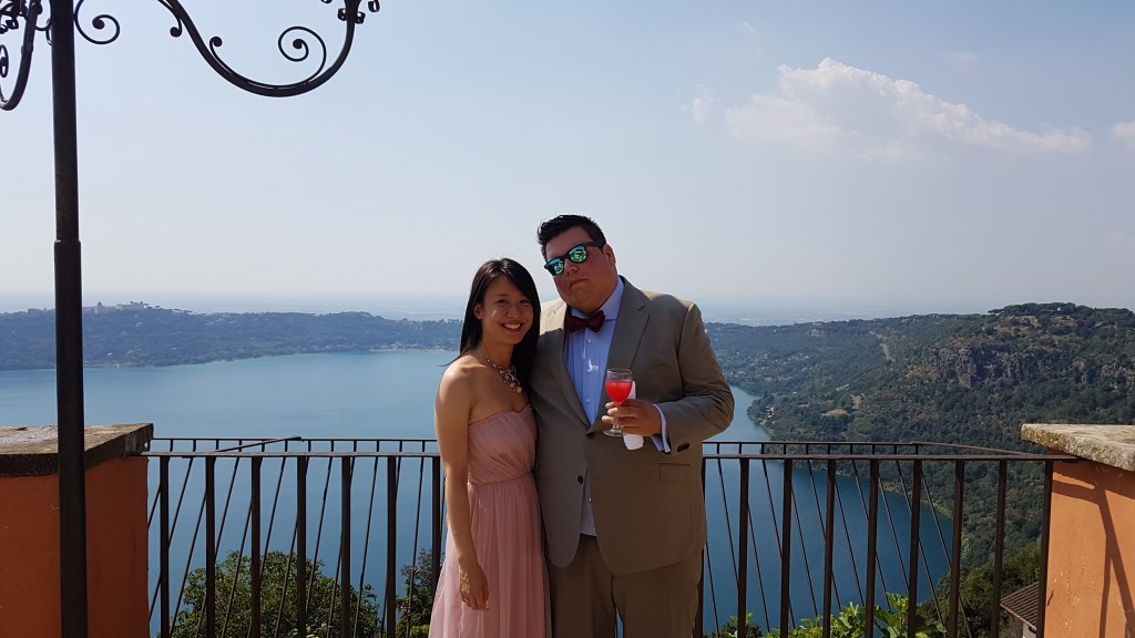 Vi en ik voor het Albanomeer (Lago Albano) op de dag van de bruiloft. Vi is een mooie roze strapless jurk. Ik in een zandkleurig pak met vlinderdas en een zonnebril op. Vi lacht mooi en ik niet.