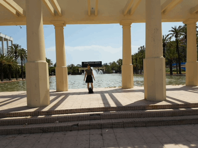 Vi in het park tussen kolommen, met palmbonen en een fontein op de achtergrond.