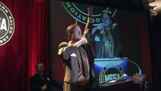 Dwight Schrute uit the Office US. Episode Dwight's Speecht. Still tijdens de speech van Dwight op het podium voor publiek, met gebalde vuist en z'n eigen beeld op een scherm achterhem.