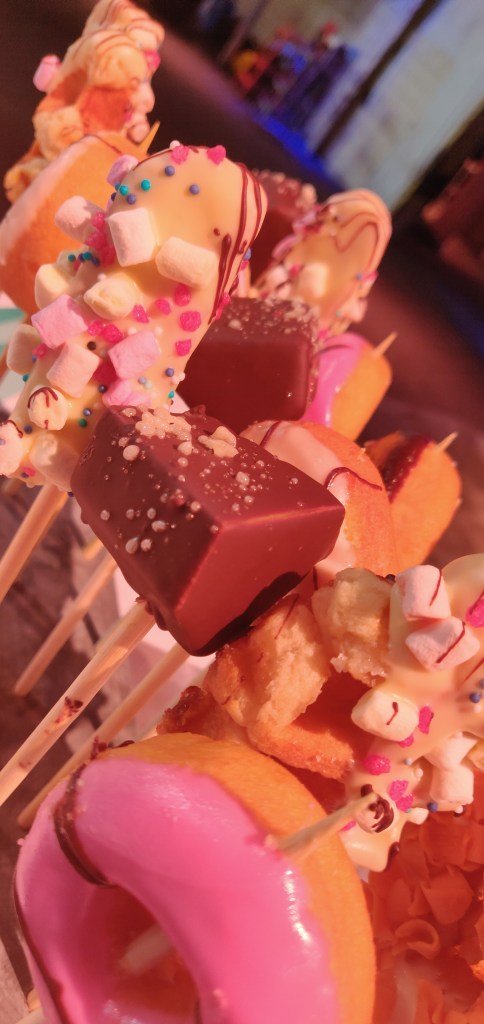 Cake lolly's, op de QX Day wordt goed voor je gezorgd. Donuts, brownies en andere cakejes op een stokje, vrolijk versier met verschillende kleuren glazuur, mini marshmellows en ander suikergoed.