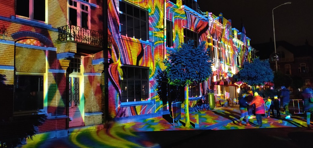 Meerdere huizen en mensen in het werk Diving in the Sea Colors. Een plein waarbij verschillende kleuren licht op geprojecteerd zijn.