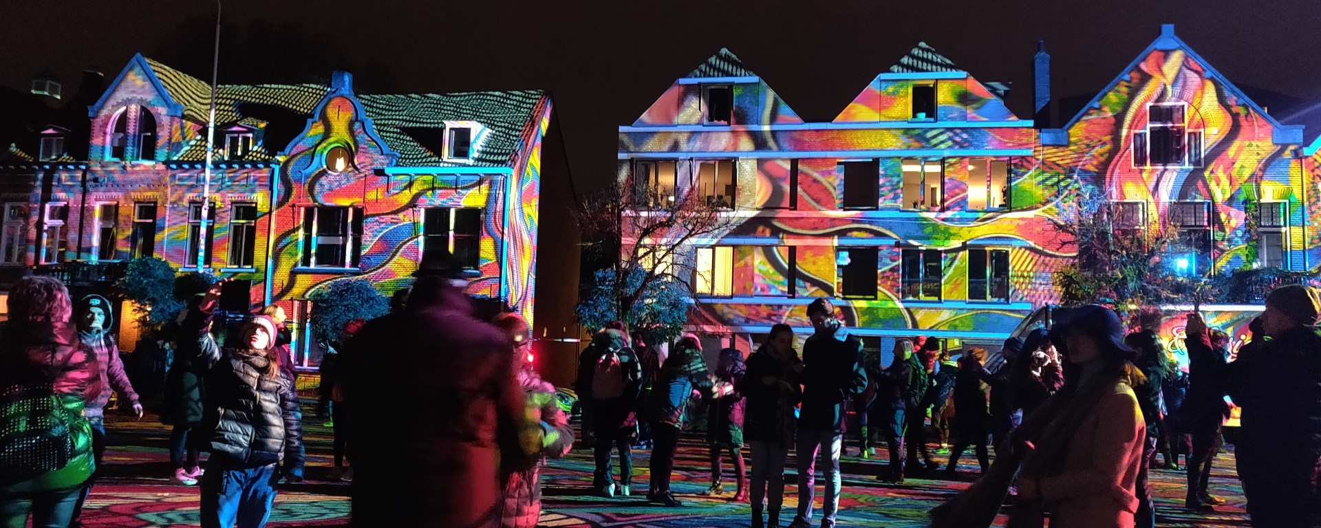 Meerdere huizen en mensen in het werk Diving in the Sea Colors. Een plein waarbij verschillende kleuren licht op geprojecteerd zijn.