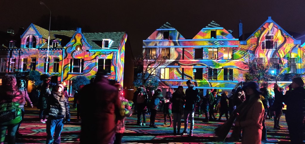 Meerdere huizen en mensen in het werk Diving in the Sea Colors. Een plein waarbij verschillende kleuren licht op geprojecteerd zijn.