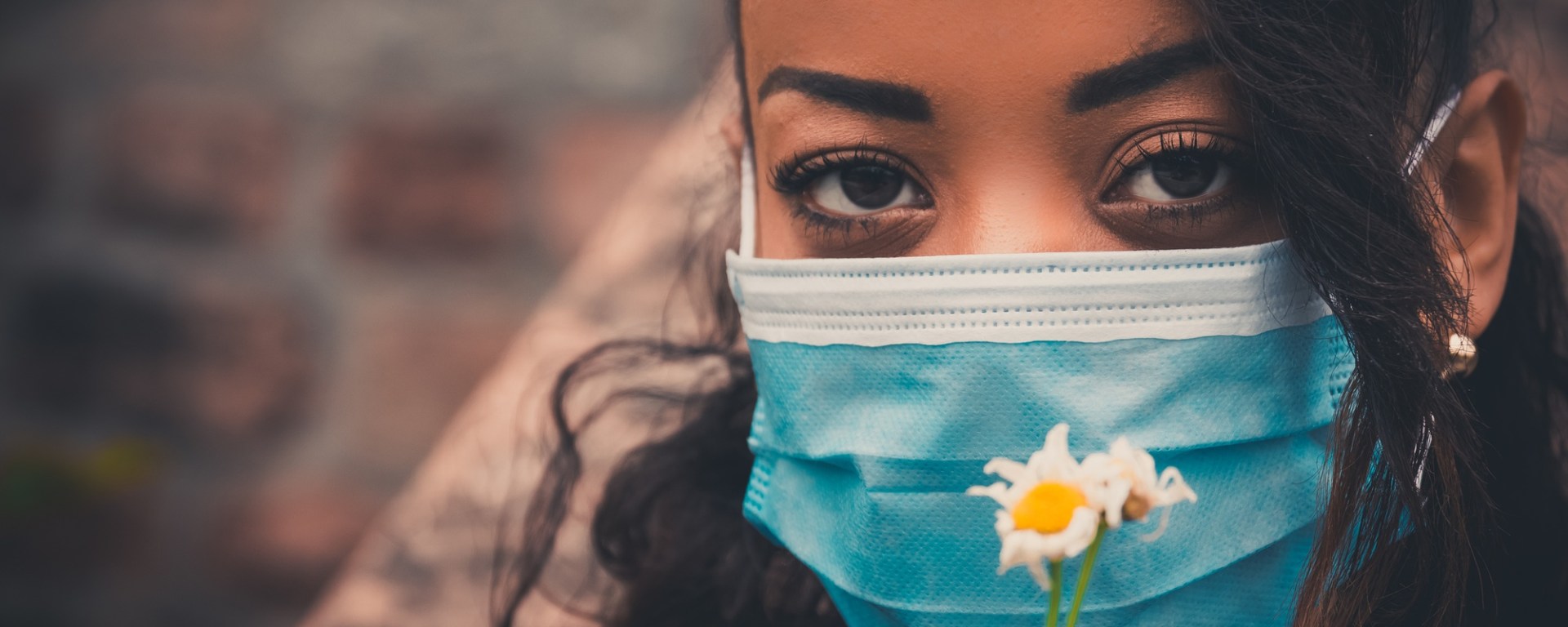 Vrouw met een blauw medisch masker kijkt indringend in de camera. Ze houd kleine wit-gele bloemen voor haar masker tegen een vage achtergrond.