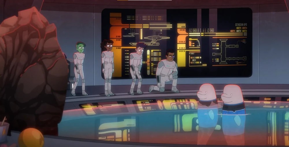 Uit de animatieserie Star Trek: Lower Decks: Cetacean Ops op de Cerritos, met een zwembad met beloega's, lieutenants Kimolu en Matt in blauwe Starfleet-uniformen met hun hoofden uit het water stekend. Naast het bad in ruimtepakken staan ​Ensigns Tendi, een Orion, Rutherford, een mens met cyborgimplantaten, en Boimler en Mariner, beiden mensen. Rutherford zit op één knie en kijkt serieus. De anderen staan ​​en kijken bezorgd. Achter de ensigns staan ​​grote LCARS-schermen (ook bekend als Okudagrams) met schema's van het schip. De LCARS weerspiegelen in het badwater.