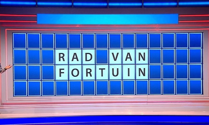 Rad van Fortuin bord uit de oude spelshow, met daarop gespeld RAD VAN FORTUIN. Er staat een man naast met de onderarmen opgeheven voorbij de ellebogen.