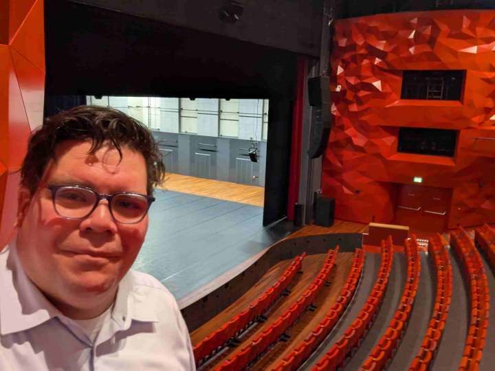 Een selfie met Randy op de voorgrond vanaf het balkon van de grote zaal in het Theater Zuidplein. Op de achtergrond het lege podium, de rode stoelen en de rode wand met de kenmerkende facetten.