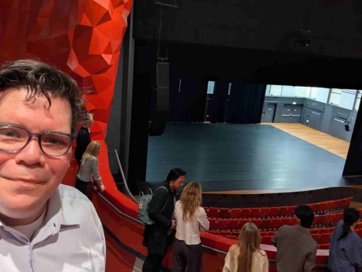 Een selfie met Randy op de voorgrond vanaf het balkon van de grote zaal in het Theater Zuidplein. Op de achtergrond het lege podium, de rode stoelen en de rode wand met de kenmerkende facetten.